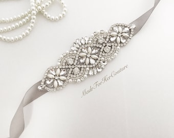 May include: A gray satin ribbon with a silver and crystal embellishment. The embellishment features a floral design with pearls and rhinestones. The text "Made For Her Couture" is printed on the ribbon.