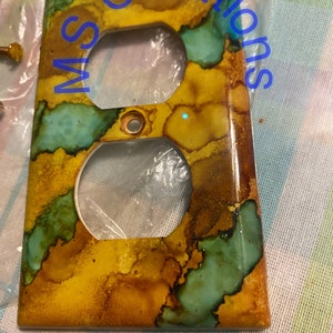 May include: A decorative electrical outlet cover with a unique, abstract design. The cover features a blend of yellow, brown, and teal colors, creating a marbled effect. The text "MS Creations" is visible in blue. The cover has two circular openings.