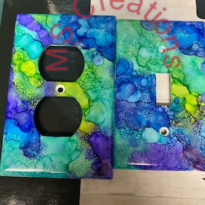 May include: Two decorative outlet covers with a blue, green, and purple abstract watercolor design. The outlet covers are made of ceramic and have a glossy finish.