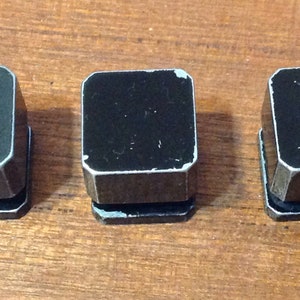 Peut inclure: Trois petits objets noirs de forme carrée avec des bordures argentées sont disposés sur une surface en bois. Les objets semblent être en métal et présentent un aspect légèrement usé. L'arrière-plan est un grain de bois brun chaud.