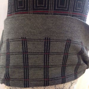 May include: A gray sweater with a geometric pattern. The top section features a black and gray square design, while the lower section has a gray plaid pattern with black lines and red accents. The sweater has a ribbed waistband.