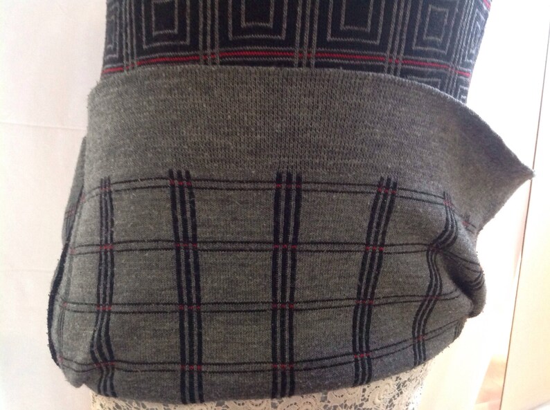 May include: A gray sweater with a geometric pattern. The top section features a black and gray square design, while the lower section has a gray plaid pattern with black lines and red accents. The sweater has a ribbed waistband.