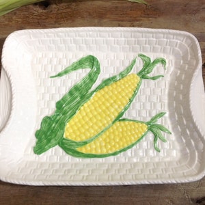 Puede incluir: Plato rectangular blanco con textura de cesta tejida. El plato presenta un diseño pintado a mano de dos mazorcas de maíz con granos amarillos y hojas verdes. Los bordes tienen un borde en forma de cuerda.