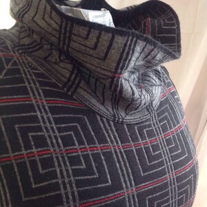 May include: A gray and black patterned turtleneck sweater with red accent lines. The sweater has a geometric design and a high, folded collar. The fabric appears to be a knit material, and the overall style is modern and warm.
