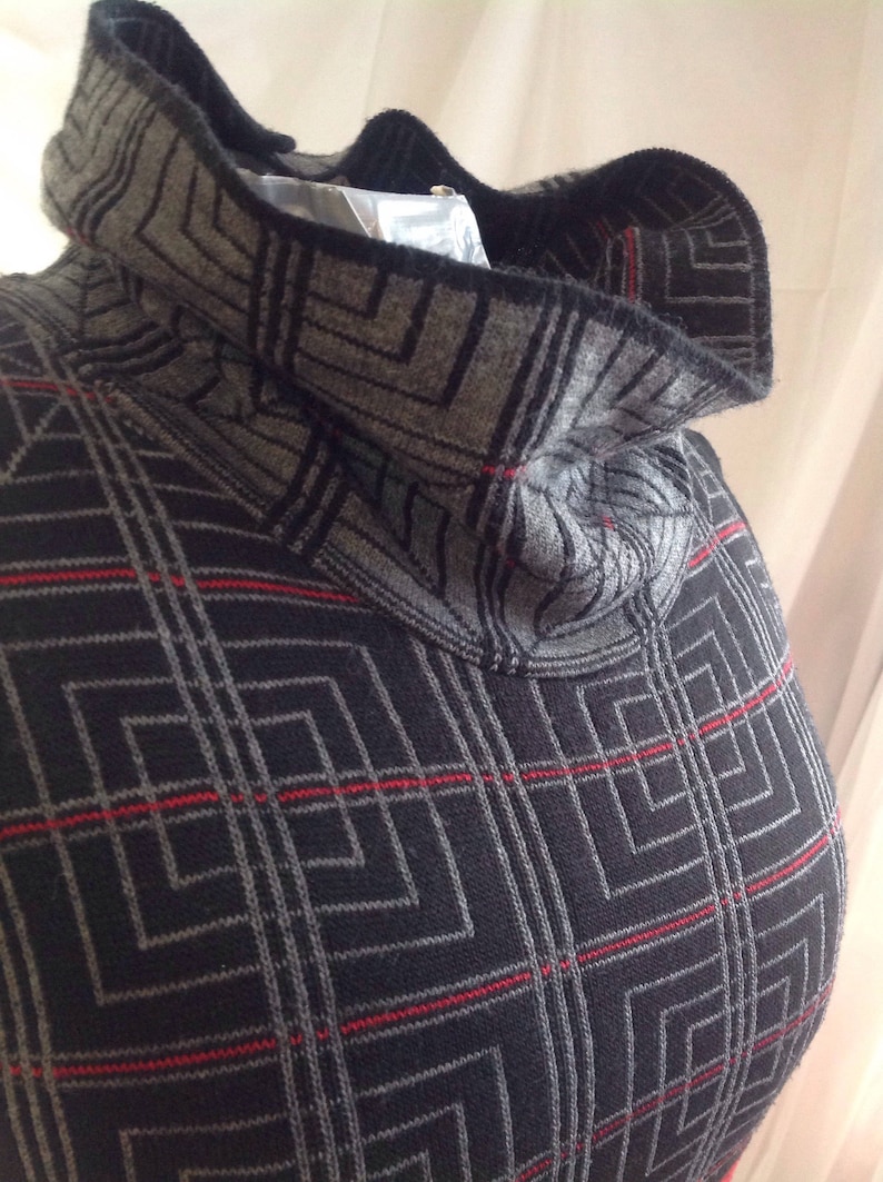 May include: A gray and black patterned turtleneck sweater with red accent lines. The sweater has a geometric design and a high, folded collar. The fabric appears to be a knit material, and the overall style is modern and warm.