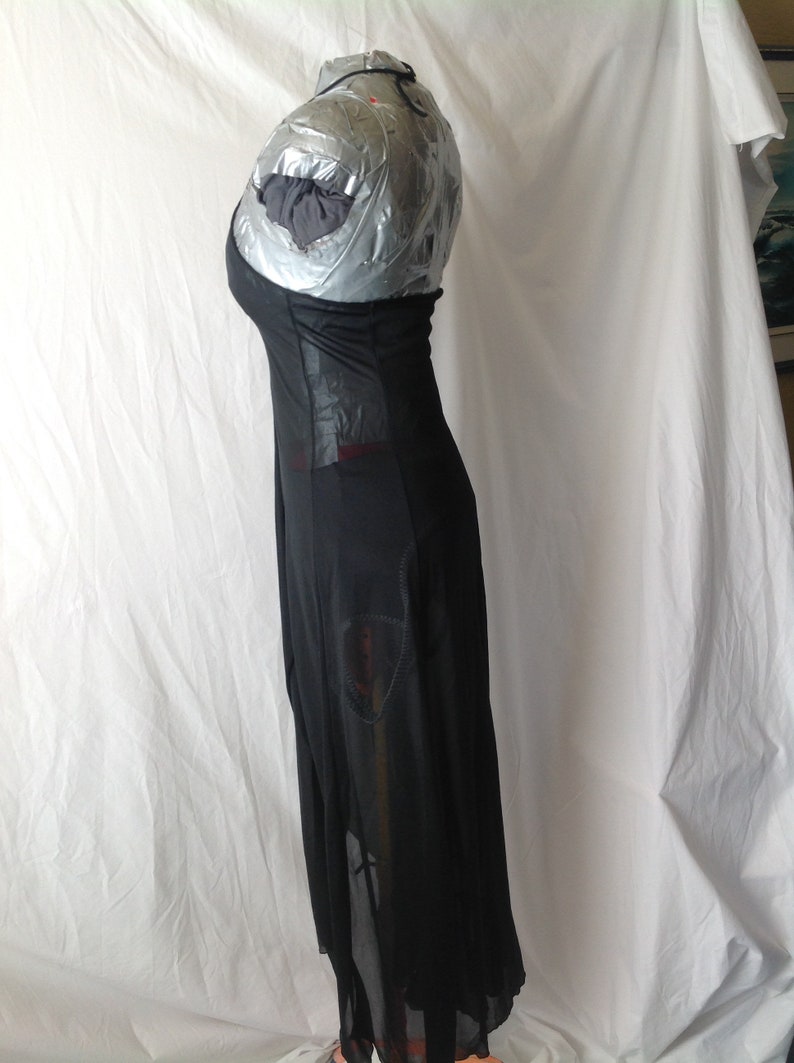 May include: A long, black dress with a sheer skirt and a fitted bodice. The dress has a unique design with cutouts and a flowing silhouette. The dress is displayed on a mannequin covered in silver tape.