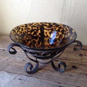 Könnte beinhalten: Eine braune Glasschale mit Leopardenmuster steht auf einem schwarzen Metallständer mit drei Beinen. Der Ständer hat ein dekoratives Rankendesign.
