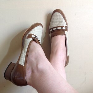 May include: A pair of white and brown heeled loafers with a perforated design. The loafers have a brown leather heel and a white leather upper with a brown leather strap across the top.