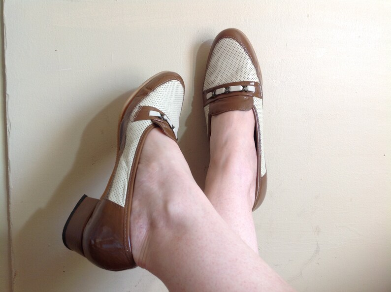 May include: A pair of white and brown heeled loafers with a perforated design. The loafers have a brown leather heel and a white leather upper with a brown leather strap across the top.