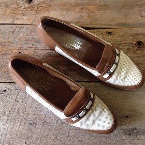 May include: A pair of brown and white leather loafers with a perforated design and a silver buckle detail. The shoes have the text "C.Brite" on the inside.