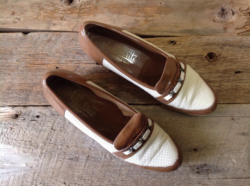 May include: A pair of brown and white leather loafers with a perforated design and a silver buckle detail. The shoes have the text "C.Brite" on the inside.