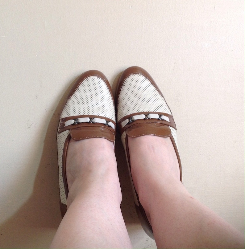May include: A pair of white and brown leather loafers with a perforated design and a row of silver embellishments on the front.