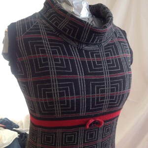 May include: A sleeveless black dress with a geometric pattern of gray squares and red lines. The dress has a high, turtleneck collar and a red band at the waist with a small bow detail. The dress is displayed on a mannequin.