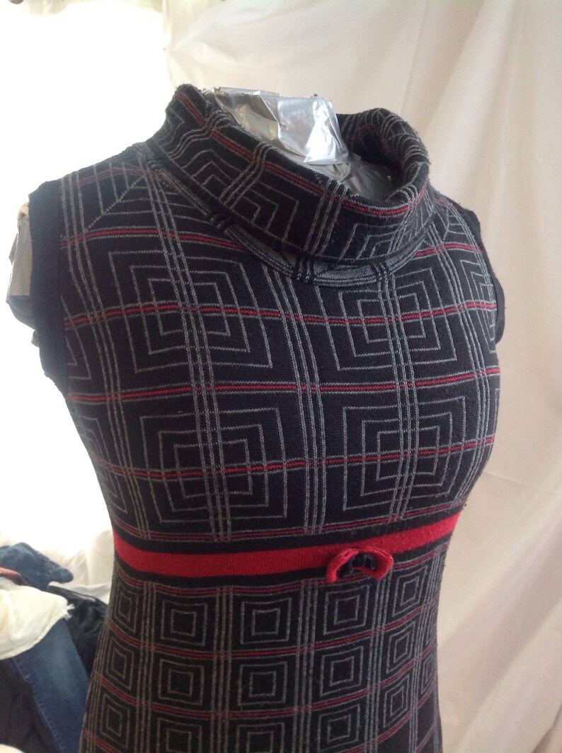 May include: A sleeveless black dress with a geometric pattern of gray squares and red lines. The dress has a high, turtleneck collar and a red band at the waist with a small bow detail. The dress is displayed on a mannequin.