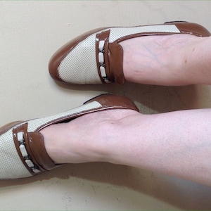 May include: A pair of white and brown leather loafers with a perforated design and a brown leather strap with a buckle. The loafers have a pointed toe and a low heel.
