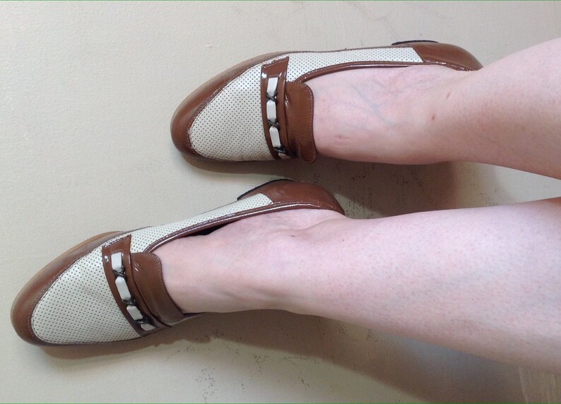 May include: A pair of white and brown leather loafers with a perforated design and a brown leather strap with a buckle. The loafers have a pointed toe and a low heel.