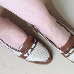 May include: A pair of white and brown leather loafers with a perforated design. The loafers have a brown leather trim and a small buckle detail.
