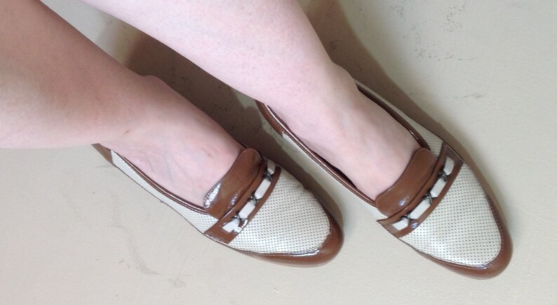 May include: A pair of white and brown leather loafers with a perforated design. The loafers have a brown leather trim and a small buckle detail.
