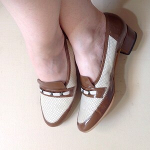 May include: A pair of brown and white leather shoes with a small block heel. The shoes have a perforated design and a white trim around the toe.
