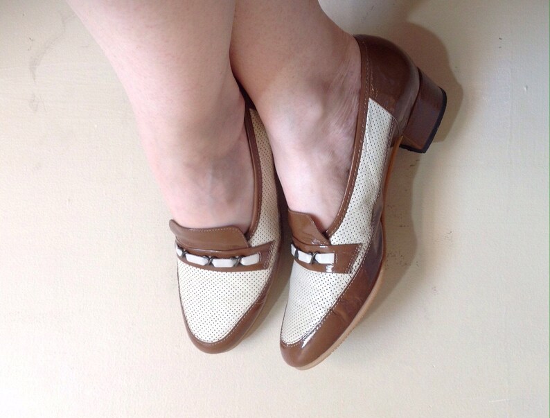 May include: A pair of brown and white leather shoes with a small block heel. The shoes have a perforated design and a white trim around the toe.