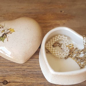 以下が含まれることがあります： 蓋に花の模様が入ったハート型の小物入れと、開いたハート型の皿。お皿にはパールのネックレスとゴールドのチェーンネックレスが入っています。木製の表面に置かれたアイテム。