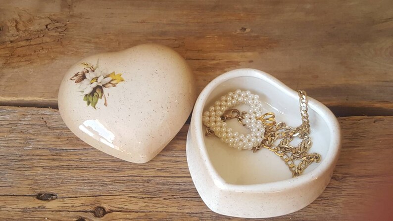 以下が含まれることがあります： 蓋に花の模様が入ったハート型の小物入れと、開いたハート型の皿。お皿にはパールのネックレスとゴールドのチェーンネックレスが入っています。木製の表面に置かれたアイテム。
