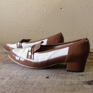 May include: A pair of brown and white heeled shoes with a perforated design. The shoes have a brown patent leather toe and heel.