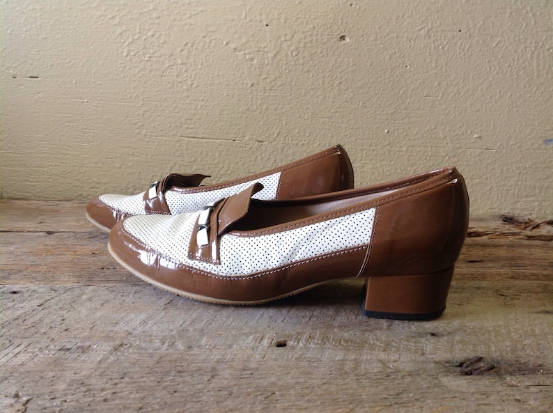 May include: A pair of brown and white heeled shoes with a perforated design. The shoes have a brown patent leather toe and heel.