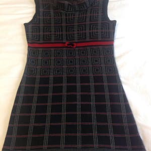 May include: Sleeveless black dress with a red ribbon and bow detail at the waist. The dress features a geometric pattern in black, red, and gray, and a turtleneck collar.