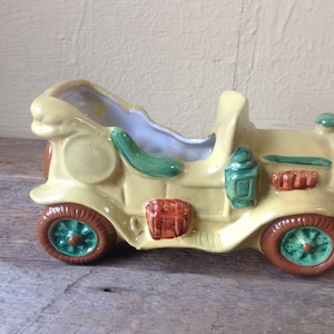 Puede incluir: Una jardinera vintage de cerámica con forma de coche en color amarillo claro. La jardinera tiene ruedas marrones con detalles verdes, detalles verdes y rojos, y un interior azul claro. El coche tiene un diseño clásico y antiguo.