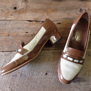 May include: A pair of brown and white leather shoes with a low heel. The shoes have a perforated design on the toe and a brown leather strap with a buckle.