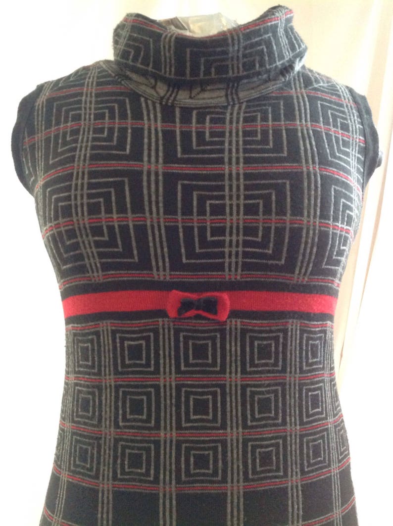May include: A sleeveless, turtleneck dress with a geometric pattern in black, gray, and red. The dress features a red band at the waist with a small bow detail. The overall design is a retro style.