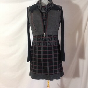 May include: A gray and black dress with a geometric pattern. The dress has long sleeves and a high neckline. A gray vest with a zipper and red stitching is layered over the dress. The dress is displayed on a mannequin.