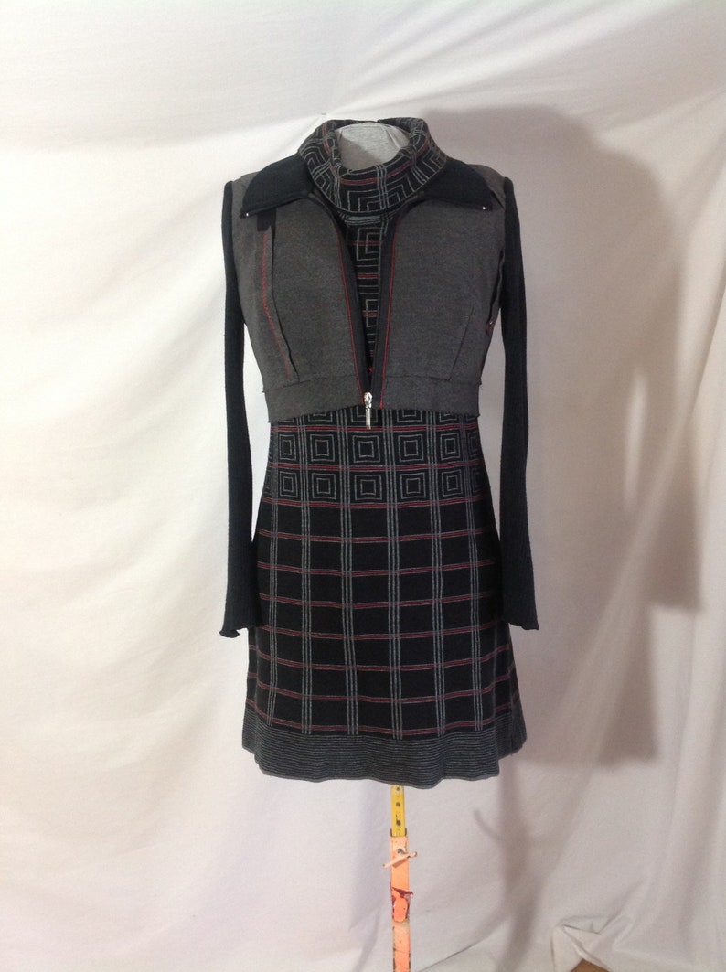May include: A gray and black dress with a geometric pattern. The dress has long sleeves and a high neckline. A gray vest with a zipper and red stitching is layered over the dress. The dress is displayed on a mannequin.