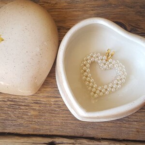 以下が含まれることがあります： 蓋に花の模様が入ったハート型の小物入れ。箱は開いており、内側には真珠のハートペンダントが入っています。箱と蓋はクリームベージュ色で、木製の表面に置かれています。
