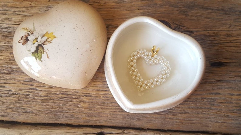 以下が含まれることがあります： 蓋に花の模様が入ったハート型の小物入れ。箱は開いており、内側には真珠のハートペンダントが入っています。箱と蓋はクリームベージュ色で、木製の表面に置かれています。