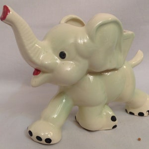 Peut inclure: Figurine d'éléphant en céramique vert clair et crème. L'éléphant a des yeux noirs et des taches noires sur les pieds. La trompe est levée et la bouche est ouverte avec un intérieur rouge. Un objet décoratif.