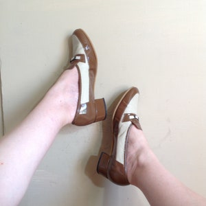 May include: A pair of brown and beige heeled shoes with a loafer style. The shoes have a brown leather upper and a beige fabric upper with a woven pattern. The heels are brown and blocky.