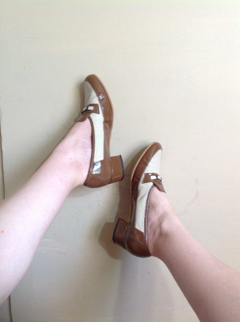 May include: A pair of brown and beige heeled shoes with a loafer style. The shoes have a brown leather upper and a beige fabric upper with a woven pattern. The heels are brown and blocky.