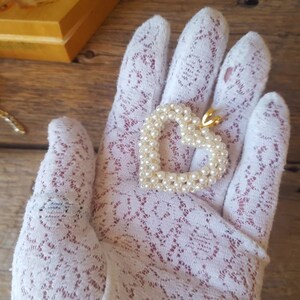 以下が含まれることがあります： 小さな白い真珠で作られたハート型のペンダント。金色のベイルが付いています。ペンダントは白いレースの手袋の上にあります。画像には、小さな装飾的な皿と木製の箱も含まれています。