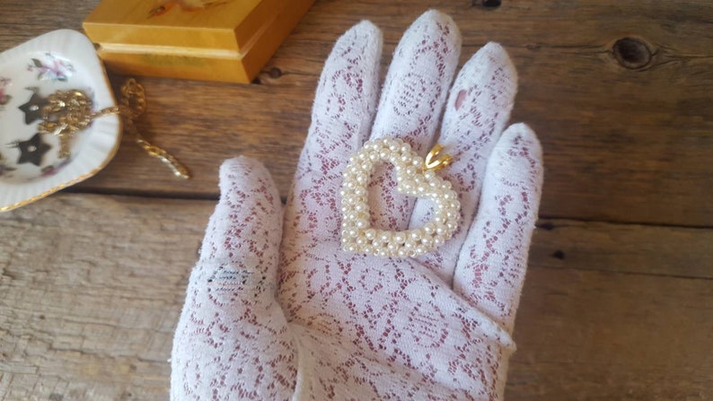 以下が含まれることがあります： 小さな白い真珠で作られたハート型のペンダント。金色のベイルが付いています。ペンダントは白いレースの手袋の上にあります。画像には、小さな装飾的な皿と木製の箱も含まれています。