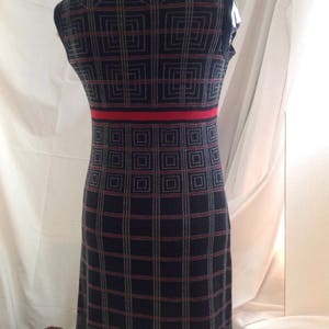 May include: A sleeveless, knee-length dress with a geometric pattern. The dress features a dark gray base with a grid of red and light gray lines. A red band accents the waist. The dress is displayed on a mannequin.