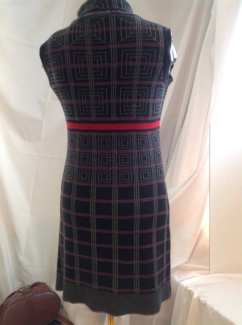 May include: A sleeveless, knee-length dress with a geometric pattern. The dress features a dark gray base with a grid of red and light gray lines. A red band accents the waist. The dress is displayed on a mannequin.