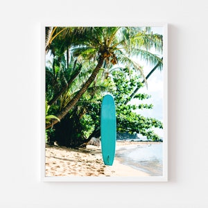 May include: A turquoise surfboard leaning against a palm tree on a sandy beach. The beach is bordered by turquoise water and lush green foliage.