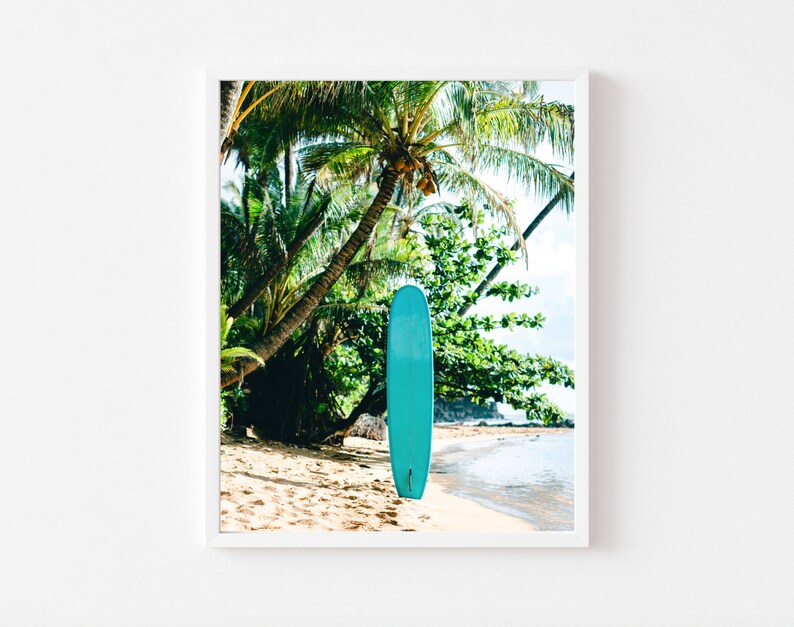 May include: A turquoise surfboard leaning against a palm tree on a sandy beach. The beach is bordered by turquoise water and lush green foliage.