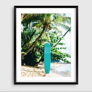 May include: A turquoise surfboard leaning against a palm tree on a sandy beach. The beach is bordered by turquoise water and lush green foliage.