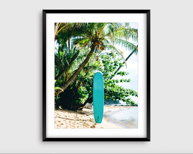 May include: A turquoise surfboard leaning against a palm tree on a sandy beach. The beach is bordered by turquoise water and lush green foliage.