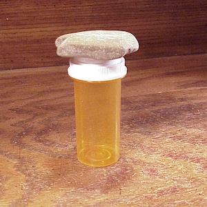 May include: An orange prescription pill bottle with a white cap, topped with a light brown, irregularly shaped stone. The bottle is empty and sits on a wooden surface.