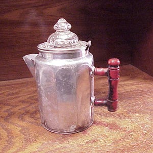 May include: A vintage aluminum percolator coffee pot featuring a cylindrical body with vertical paneling, a clear glass knob lid, and a dark red wooden handle attached with metal brackets. The spout is angled outward from the top rim.