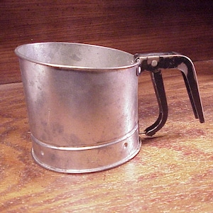 May include: A vintage-style metal flour sifter with a cylindrical body and a black handle featuring a squeeze-action trigger mechanism. The exterior of the sifter has a weathered, metallic finish with visible signs of use and a simple, functional design.
